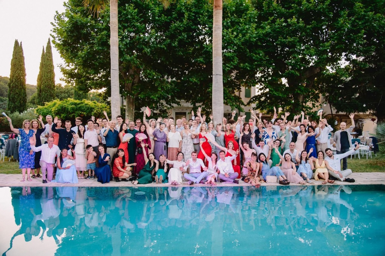 Un grand groupe de personnes pose joyeusement devant une piscine, entouré de végétation et d'arbres.