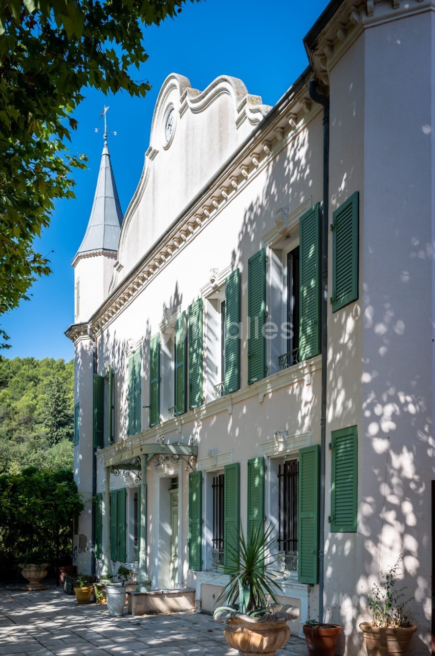 Façade d'une maison provençale blanche avec volets verts, terrasse ombragée et clocher en arrière-plan.
