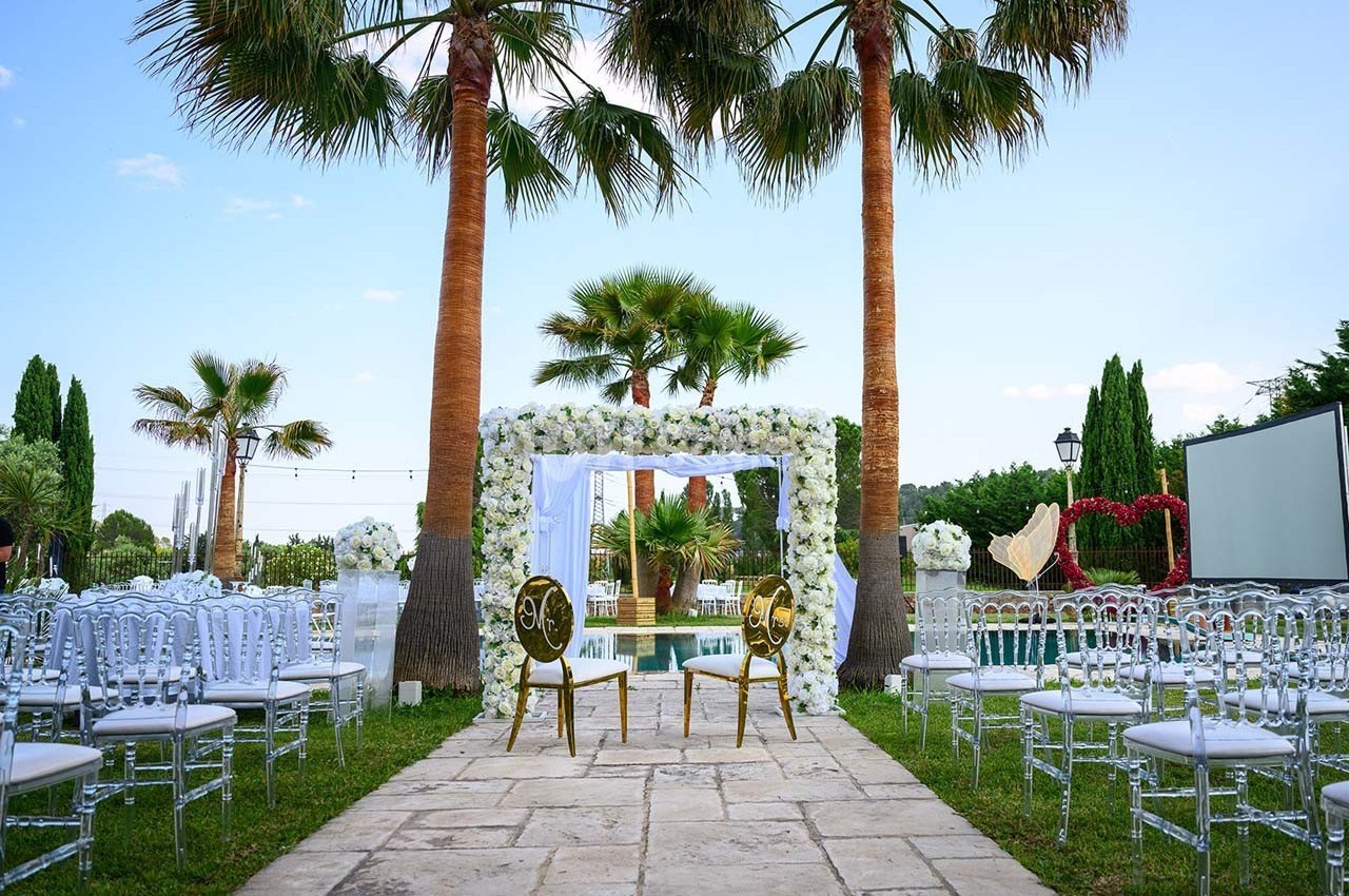 Décor de cérémonie en plein air avec arche fleurie, chaises transparentes et palmiers en arrière-plan.