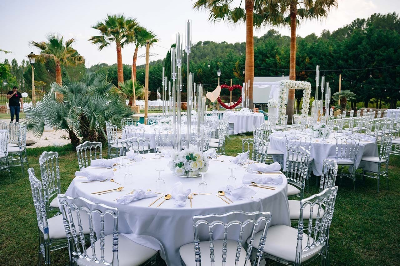 Tables rondes décorées pour un événement en plein air, avec nappes blanches, chaises transparentes et chandeliers.