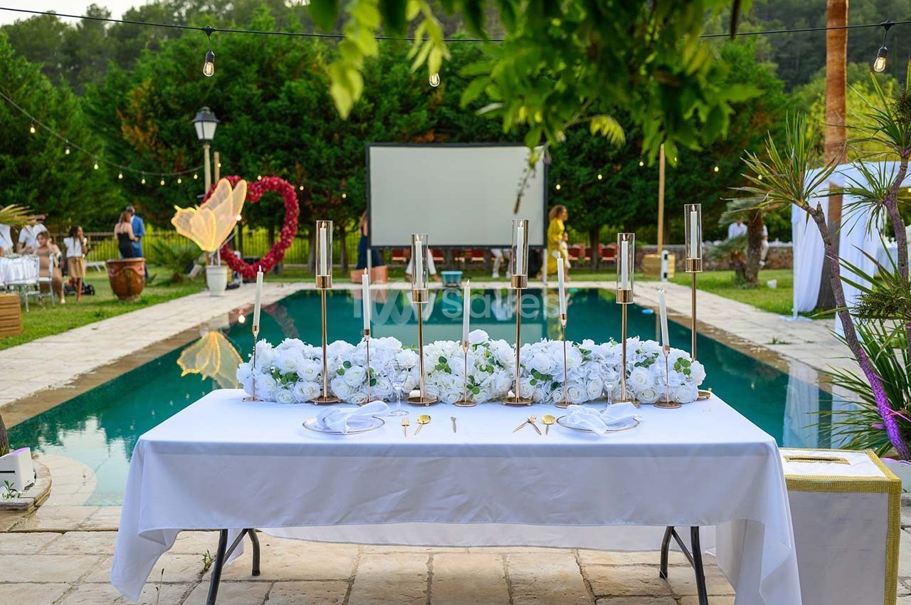 Table décorée avec des fleurs blanches et des bougies, placée devant une piscine entourée de verdure et de guirlandes…