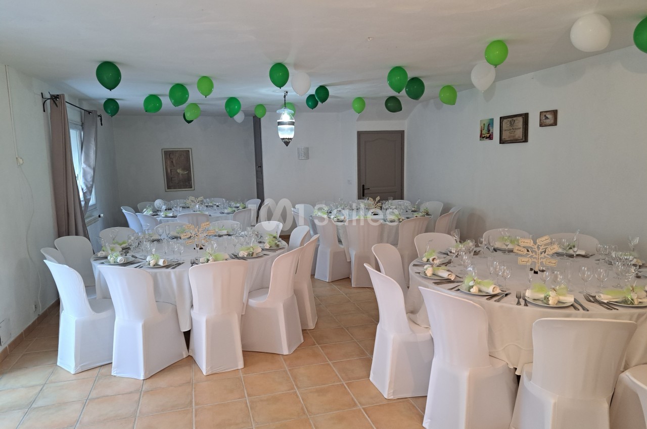 Salle décorée pour un événement avec des tables rondes dressées, nappes blanches et ballons verts suspendus.