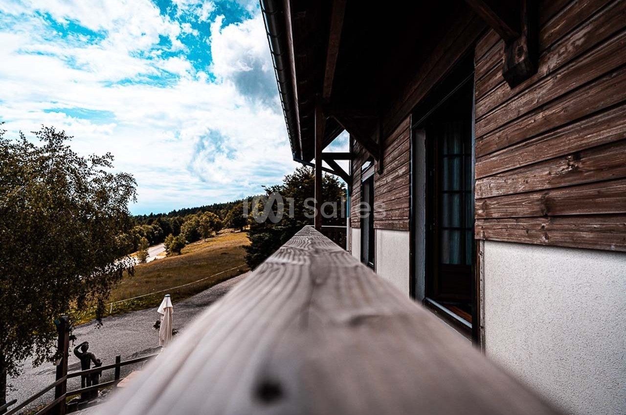 Vue depuis un balcon en bois d'une maison, donnant sur un paysage naturel avec arbres, prairie et ciel partiellement nuageux.