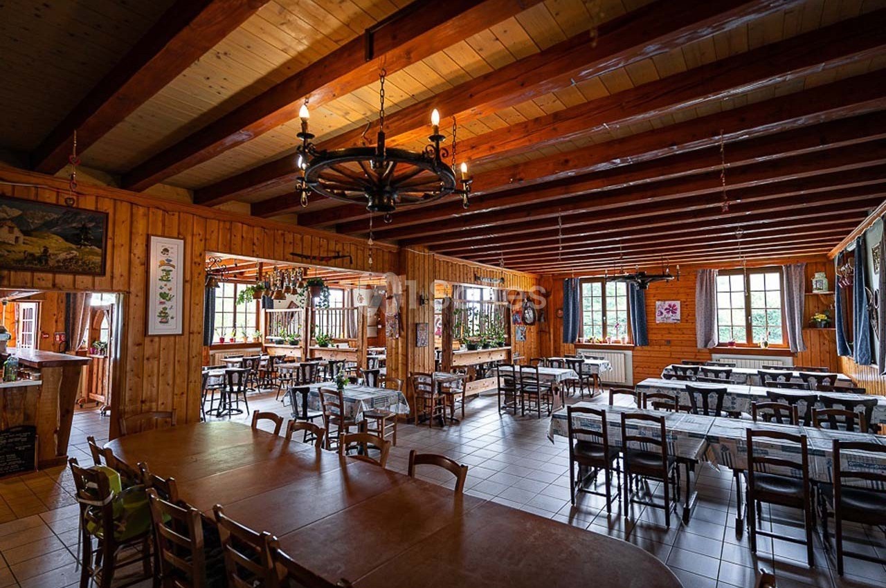 Salle à manger spacieuse en bois avec tables et chaises alignées, éclairée par des fenêtres et des lustres rustiques.