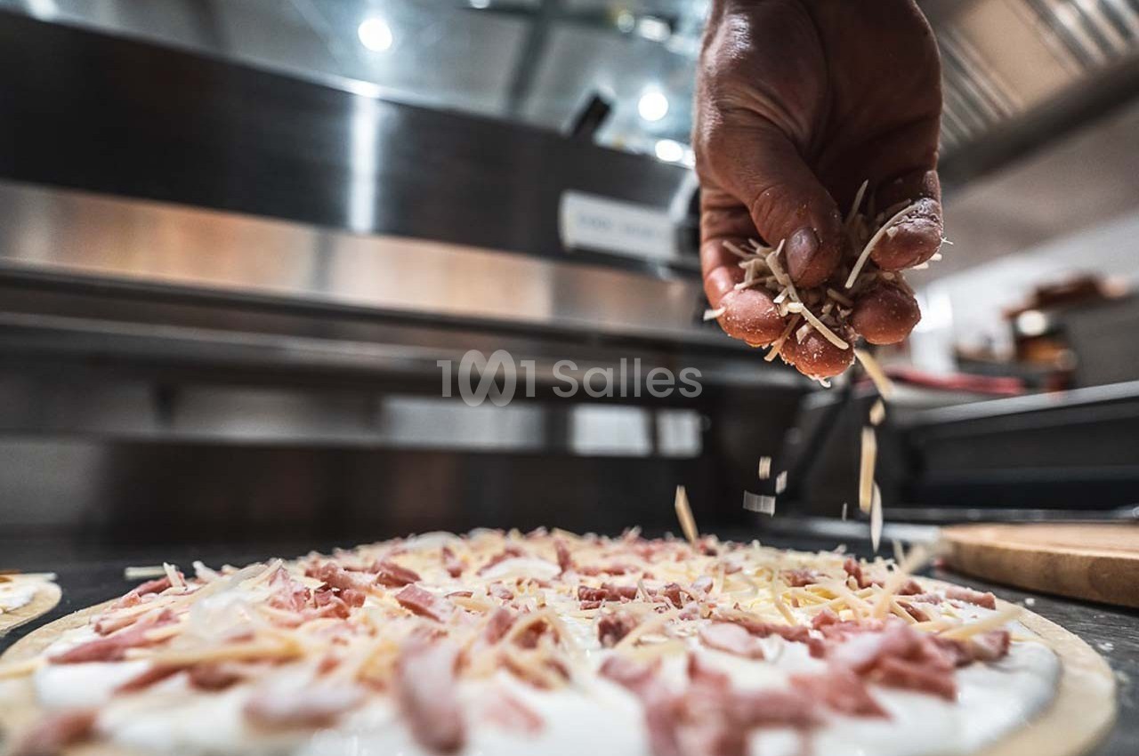 Une main saupoudre du fromage râpé sur une pizza garnie de jambon dans une cuisine professionnelle.