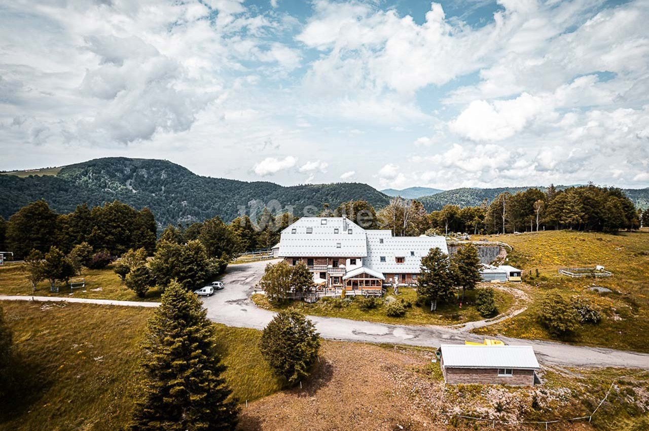 Vue aérienne d'un bâtiment entouré de prairies, d'arbres et de montagnes sous un ciel partiellement nuageux.