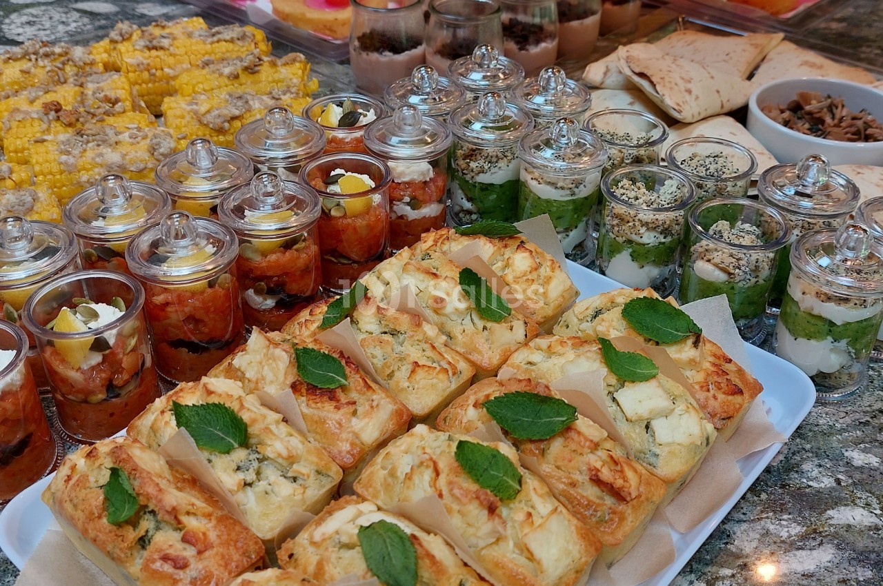Plateau de buffet avec verrines salées, épis de maïs grillés, pains garnis et accompagnements variés sur une table en pierre.