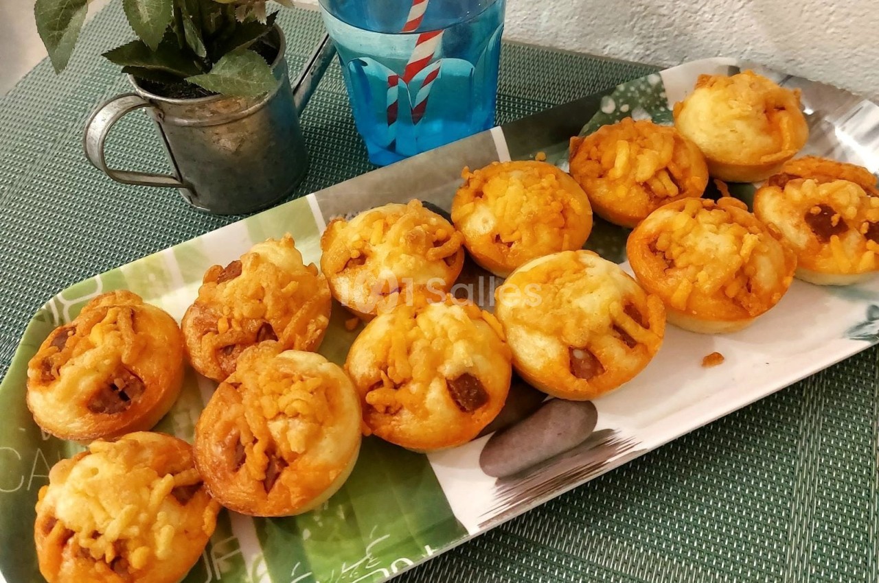Plateau de mini muffins salés dorés, disposés en rangées, sur une table avec une plante et un verre d'eau.