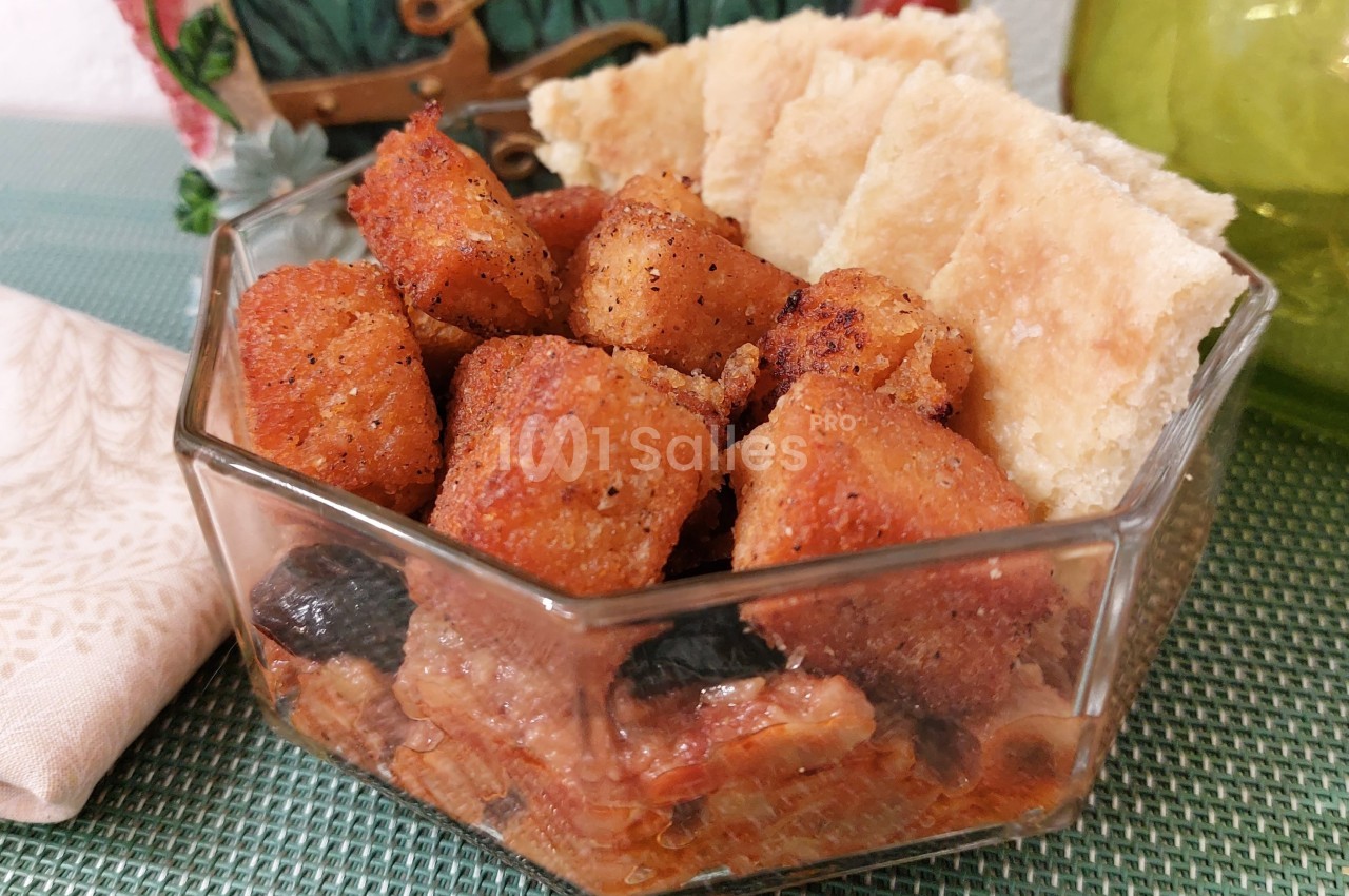 Cubes de pain frits dorés accompagnés de morceaux de pain plat et d'olives dans un bol en verre.