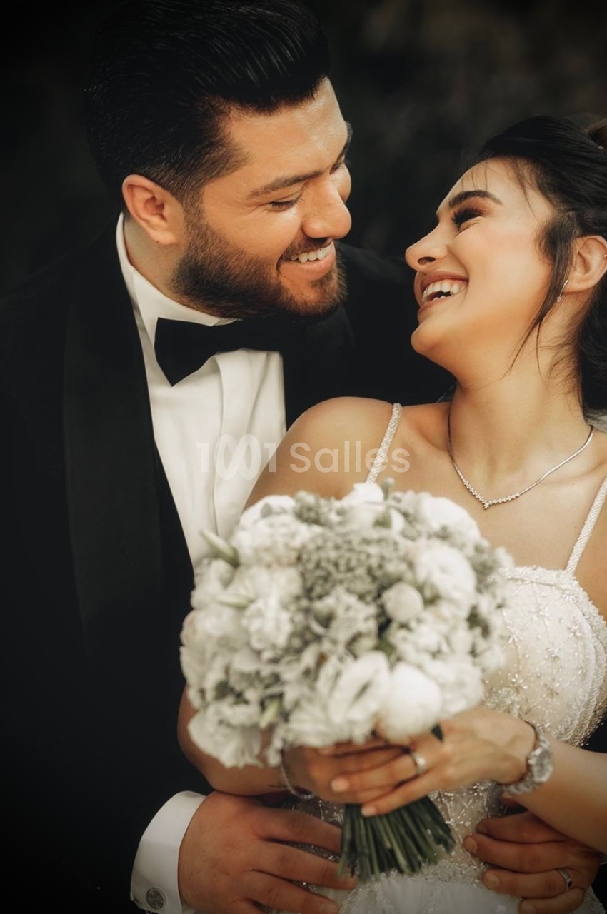 Un couple en tenue de mariage sourit et se regarde tendrement, la mariée tenant un bouquet de fleurs blanches.