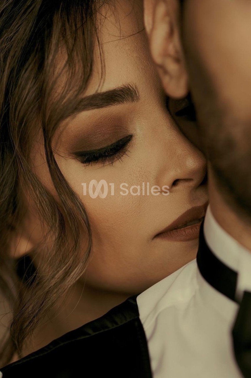 Femme au maquillage soigné posant son visage contre l'épaule d'un homme en costume, dans une ambiance intime.