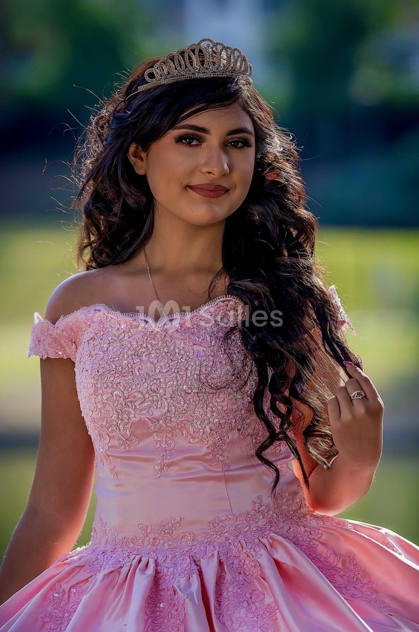 Jeune femme portant une robe rose élégante et une couronne, posant en extérieur avec un fond flou de verdure.