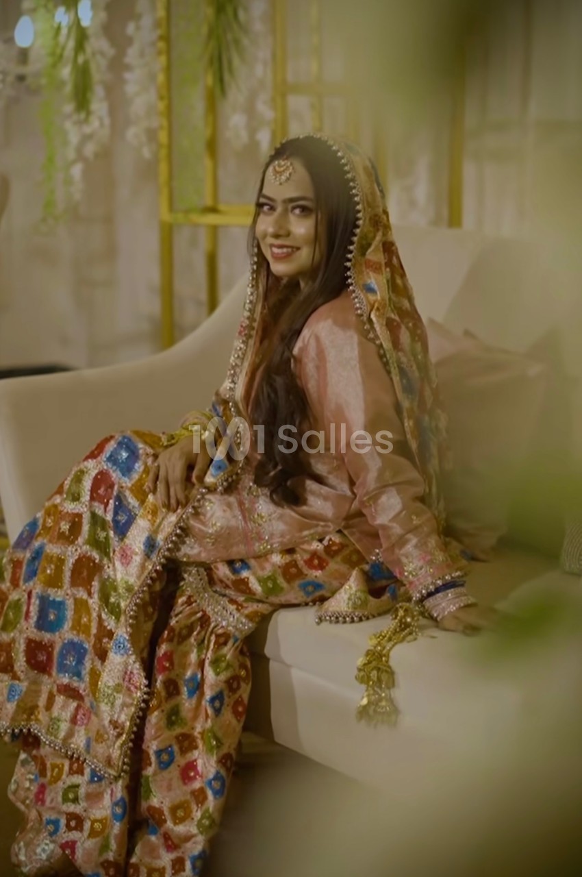 Femme souriante en tenue traditionnelle colorée, assise sur un canapé dans un décor intérieur élégant.