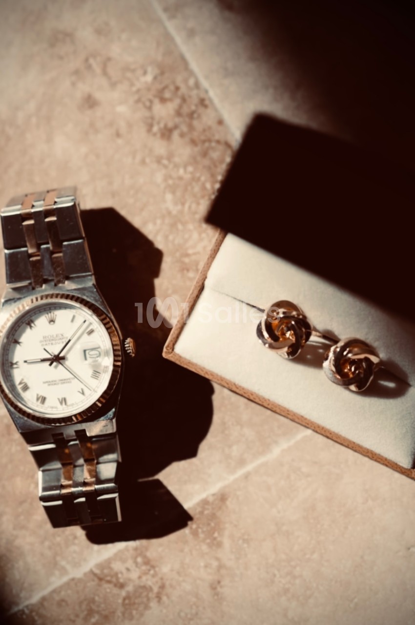 Montre en métal argenté posée sur une surface beige à côté d'une boîte contenant des boutons de manchette dorés.