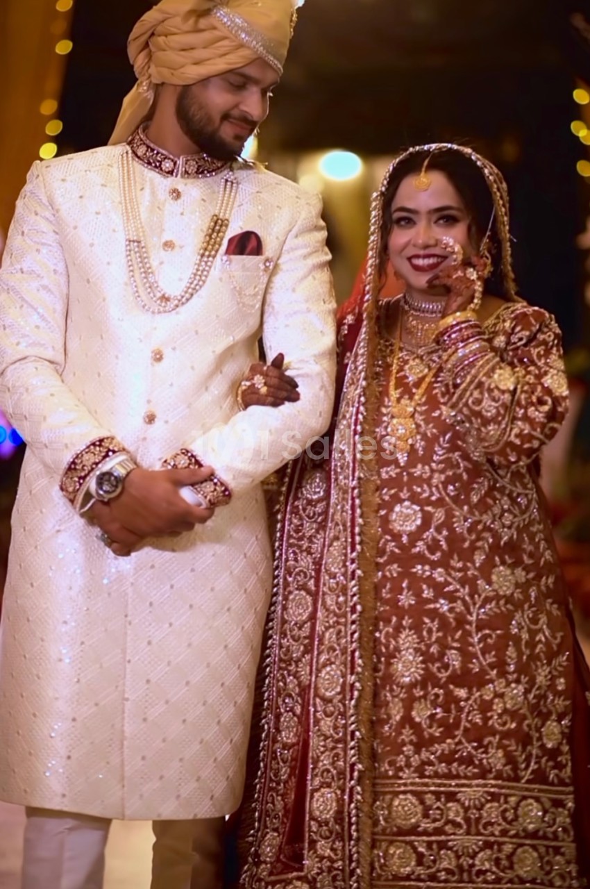 Un couple en tenue de mariage traditionnelle, l'homme en sherwani blanc et la femme en robe brodée rouge.