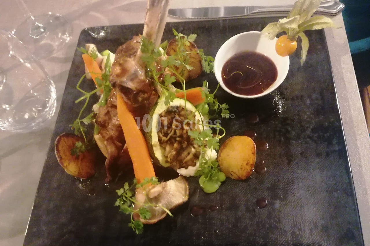 Plat gastronomique composé d'un carré d'agneau, légumes variés, sauce en bol et présentation soignée sur assiette noire.