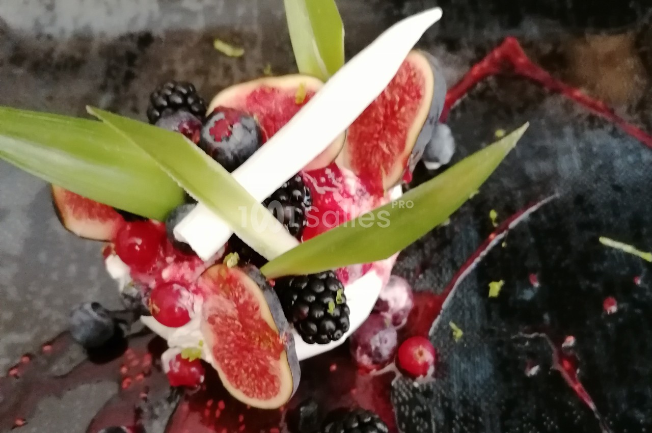 Dessert décoré de figues, mûres, groseilles, myrtilles, feuilles vertes et éclats blancs sur une assiette sombre.