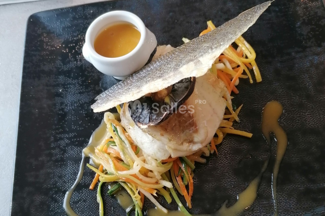 Plat de poisson avec légumes croquants, sauce servie à part dans une coupelle blanche, présenté sur une assiette noire.