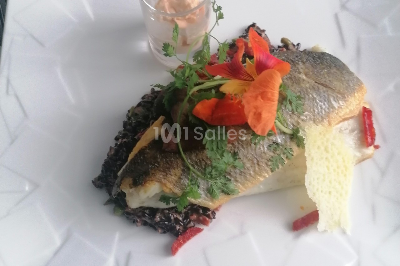 Filet de poisson grillé sur lit de riz noir, décoré de fleurs comestibles et accompagné d'une sauce en verrine.