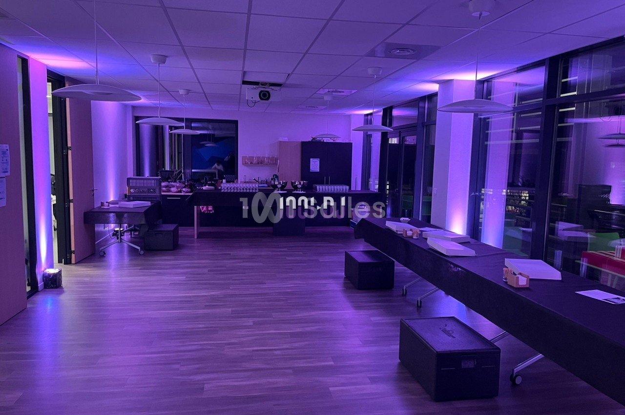 Salle moderne avec éclairage violet, tables alignées, équipement audio et grandes baies vitrées donnant sur l'extérieur.