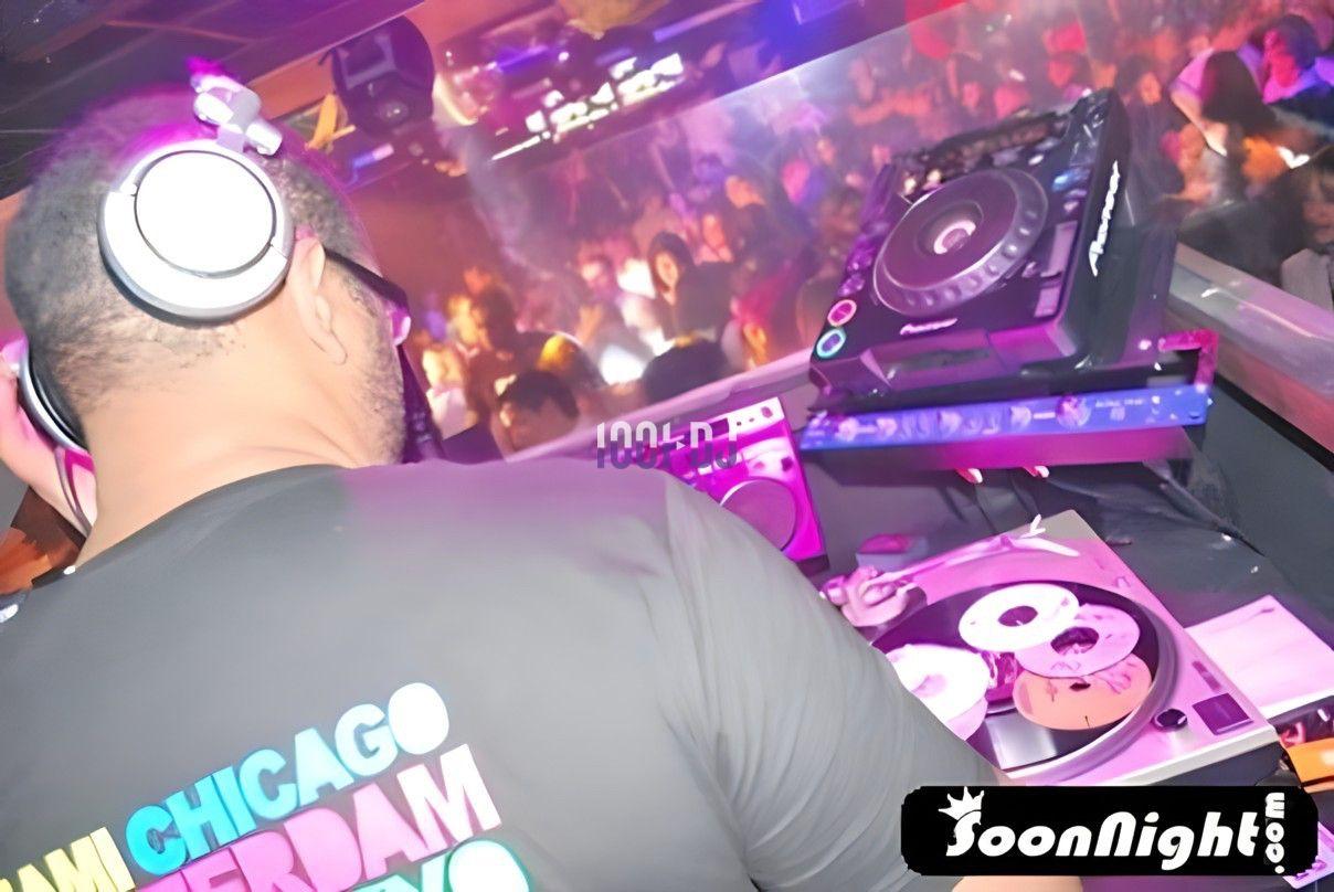Un DJ mixe sur des platines dans une boîte de nuit animée, avec une foule dansante en arrière-plan.
