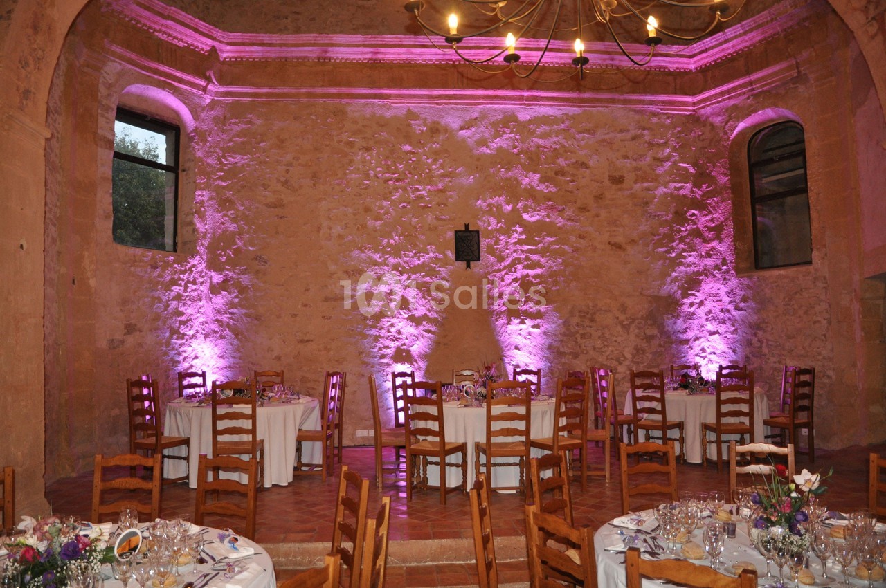 Salle en pierre avec tables dressées pour un événement, éclairée par des lumières roses et un lustre au plafond.