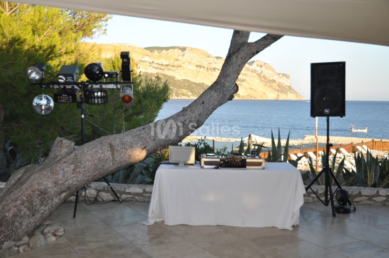 Table de mixage sous un auvent avec vue sur la mer, un arbre incliné et des équipements audio-visuels à proximité.