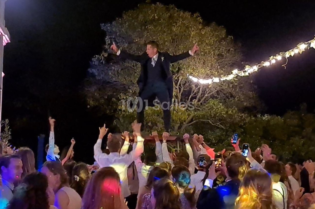 Un homme en costume est porté par une foule festive sous des guirlandes lumineuses, en extérieur de nuit.