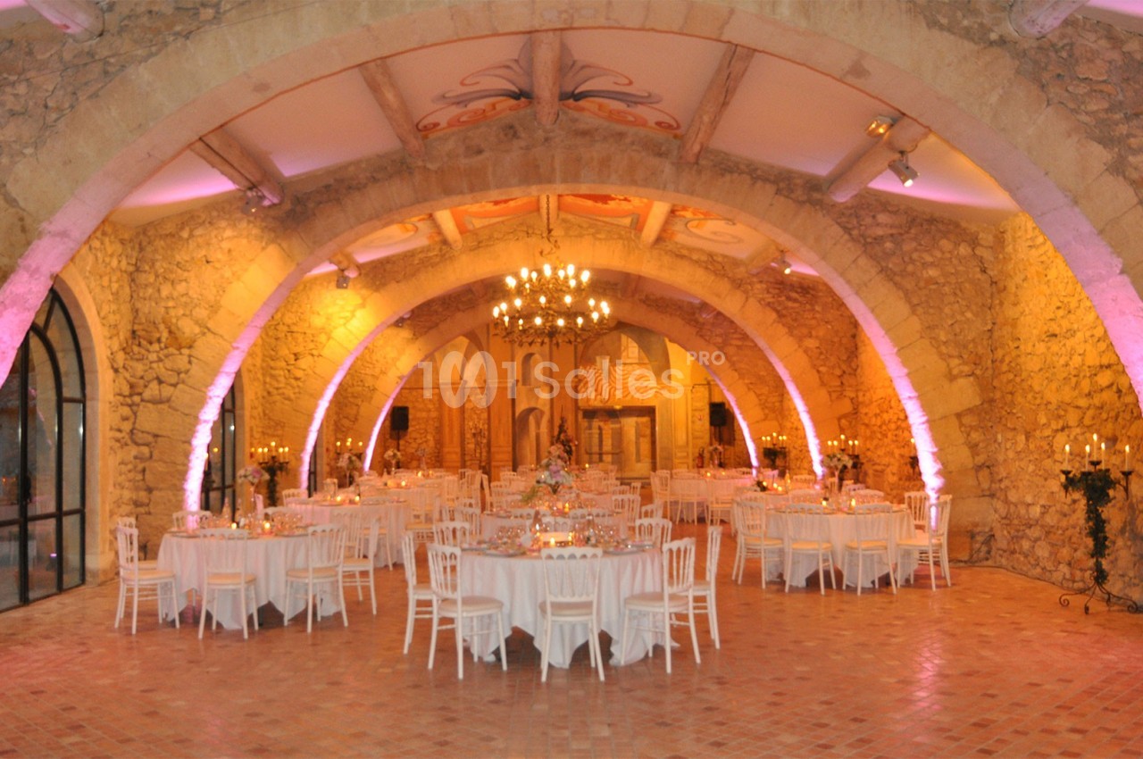 Salle voûtée en pierre avec tables rondes dressées pour un événement, éclairage chaleureux et lustre central.