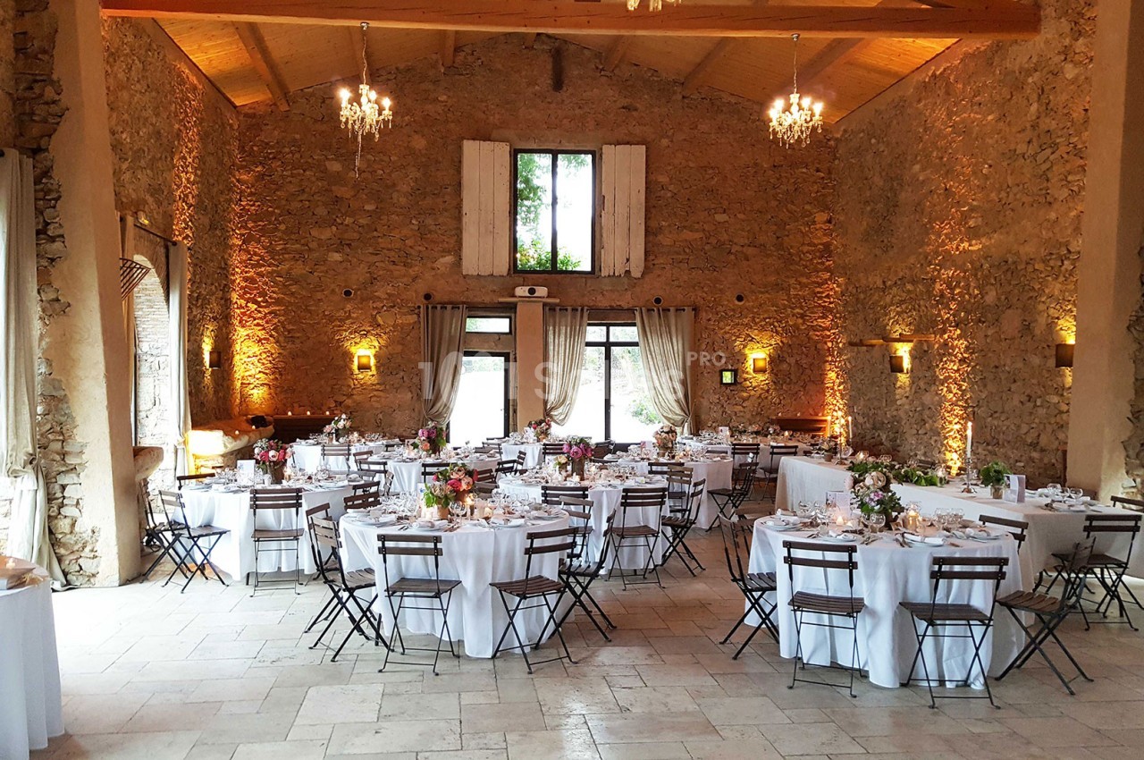 Salle de réception en pierre avec tables rondes dressées, chaises pliantes et lumière naturelle entrant par les fenêtres.