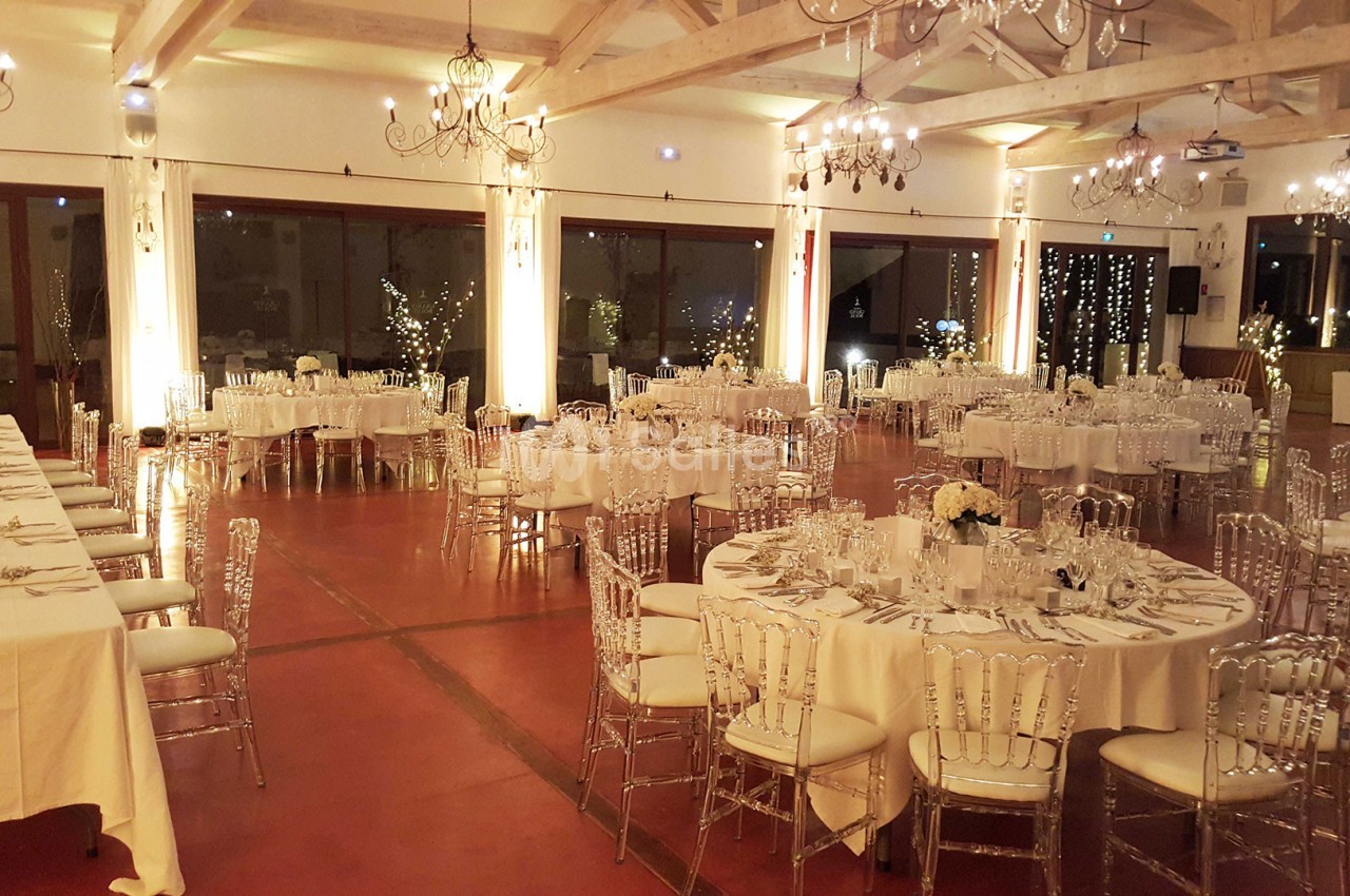 Salle de réception élégamment décorée avec des tables rondes dressées, des chaises transparentes et des lumières tamisées.