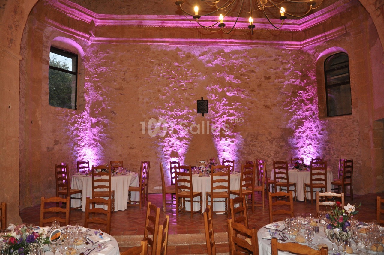 Salle en pierre avec tables dressées pour un événement, éclairée par des lumières roses et un lustre au plafond.