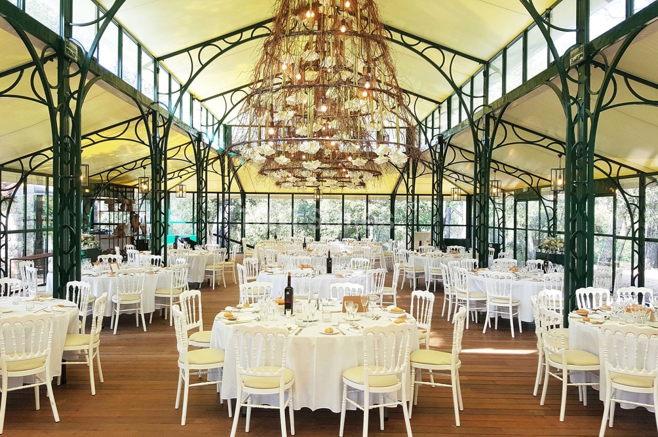 Salle de réception lumineuse avec tables rondes dressées, chaises blanches et grande suspension centrale en branches.