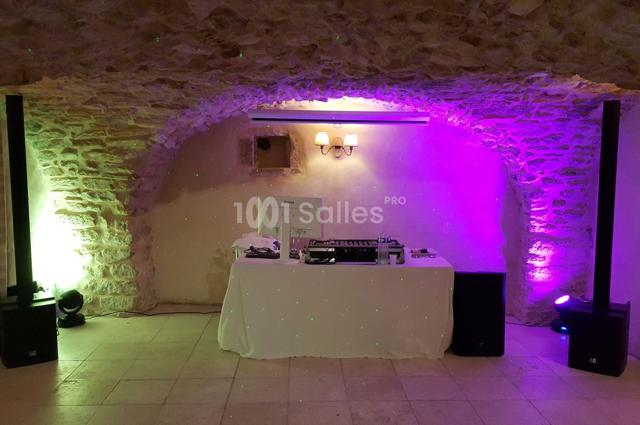 Table de DJ équipée sous une voûte en pierre, éclairée par des lumières colorées violettes et vertes.