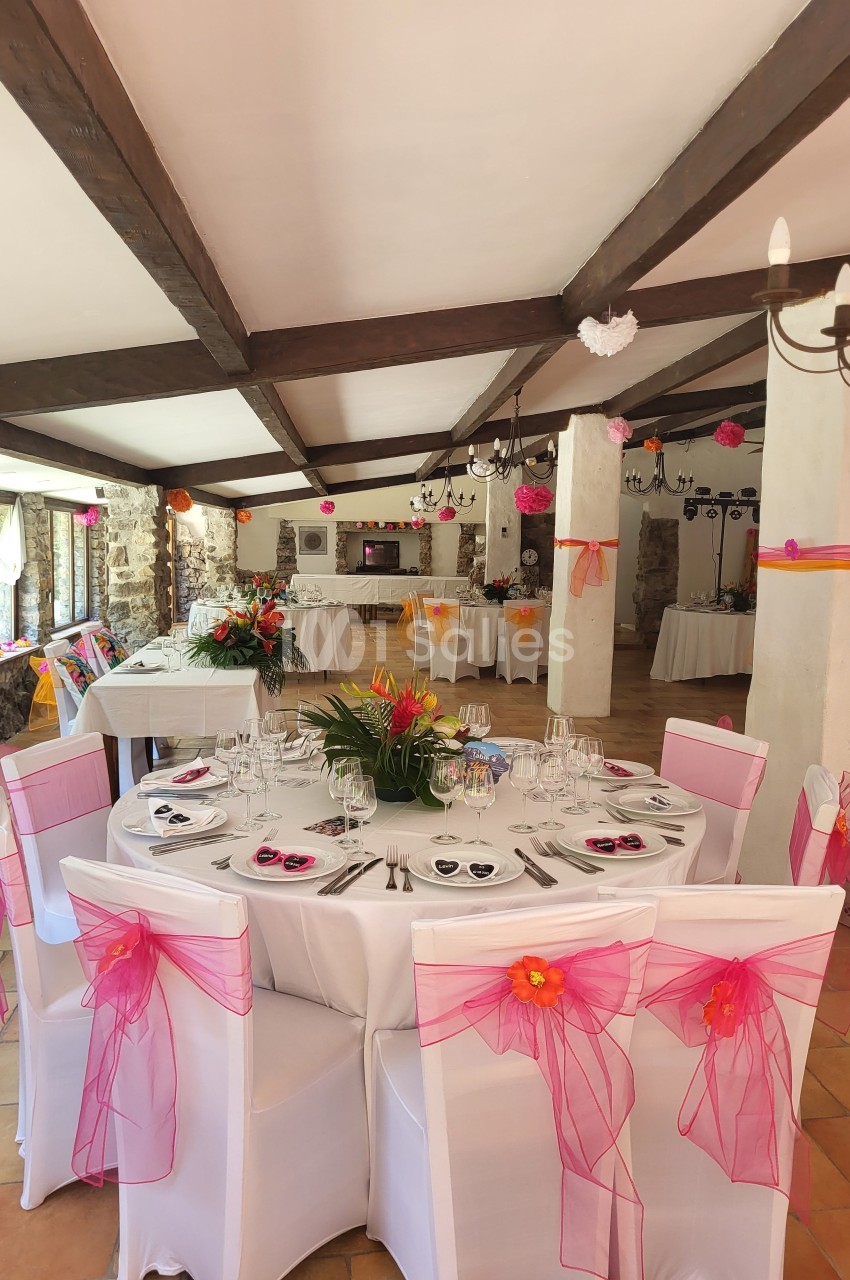Salle de réception décorée avec des tables rondes, nappes blanches et chaises ornées de nœuds roses et orange.
