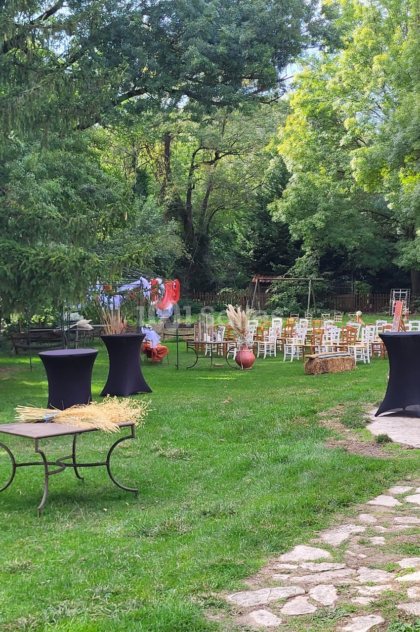 Chaises disposées en extérieur sur une pelouse entourée d'arbres, avec tables hautes décorées et bottes de paille.