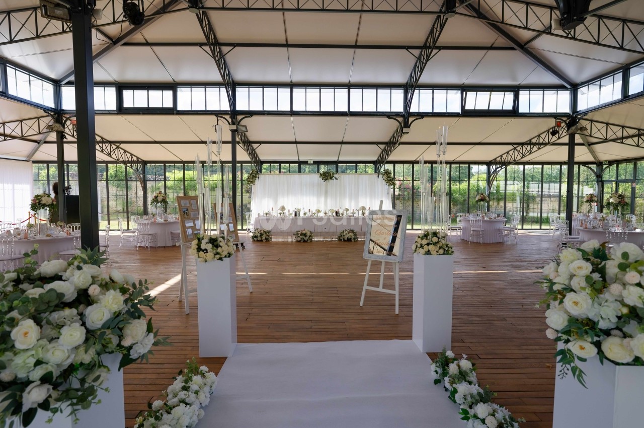 Salle de réception lumineuse avec décoration florale blanche, tables dressées et espace central en bois.