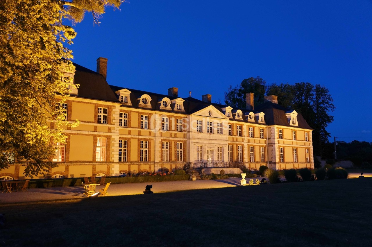 Façade éclairée d'un château historique entouré de végétation, photographiée à la tombée de la nuit.