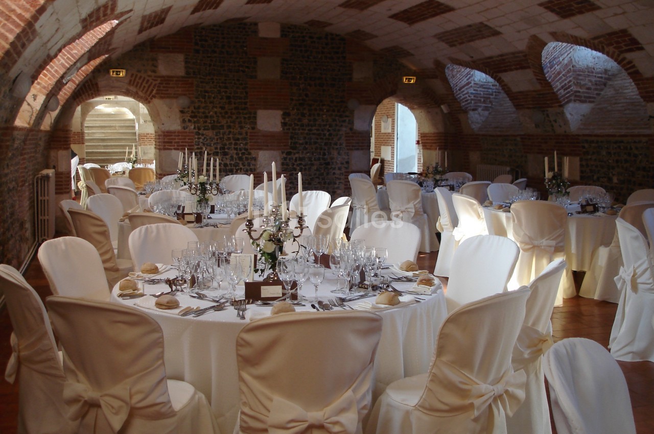 Salle voûtée en briques aménagée pour un événement avec des tables rondes dressées et des chaises couvertes de housses…