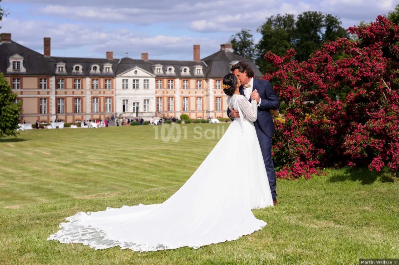 Un couple de mariés s'embrasse dans un jardin devant un château, entouré de pelouse et de fleurs rouges.
