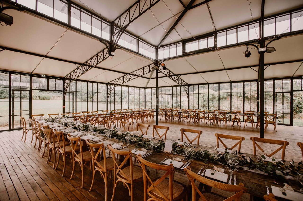 Grande salle lumineuse avec charpente métallique, tables en bois alignées et décorées de verdure, vue sur l'extérieur…