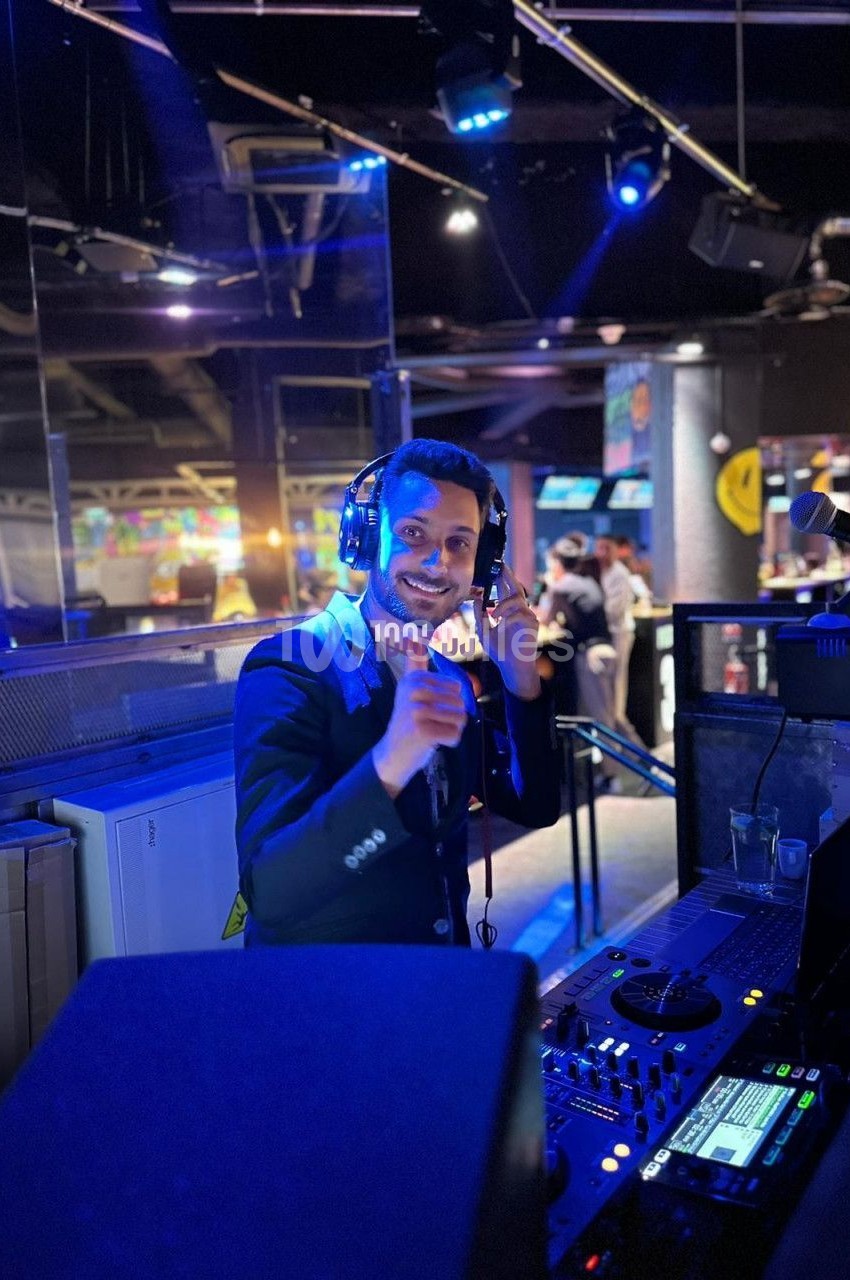 Un DJ souriant mixe de la musique dans une salle éclairée par des lumières bleues, entouré de matériel audio.