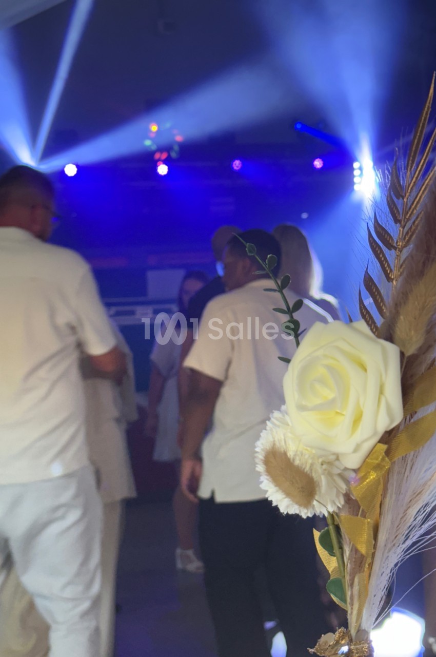 Des personnes en tenue blanche dansent sous des lumières colorées, avec un bouquet de fleurs séchées au premier plan.