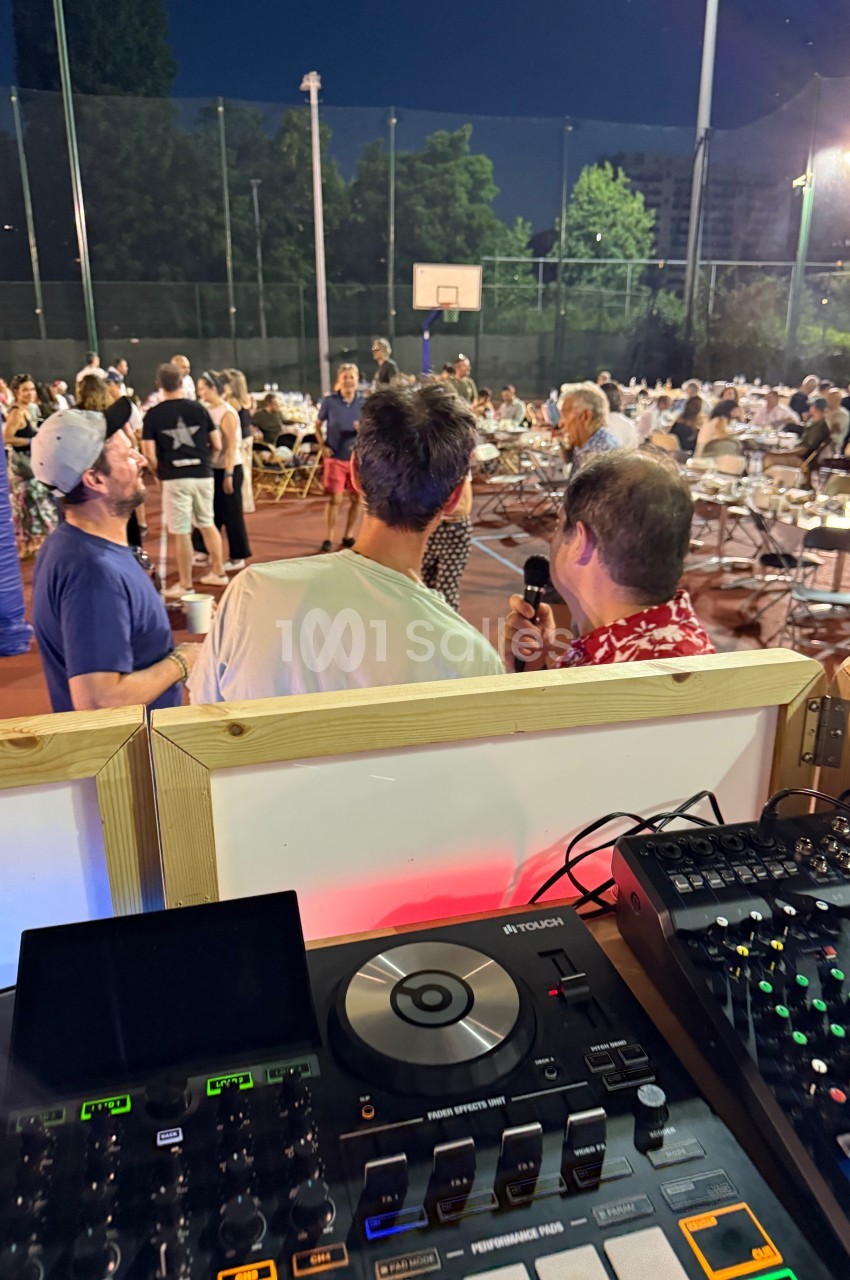 Des personnes discutent sur un terrain de sport en plein air, avec des tables installées et une console de DJ au premier…