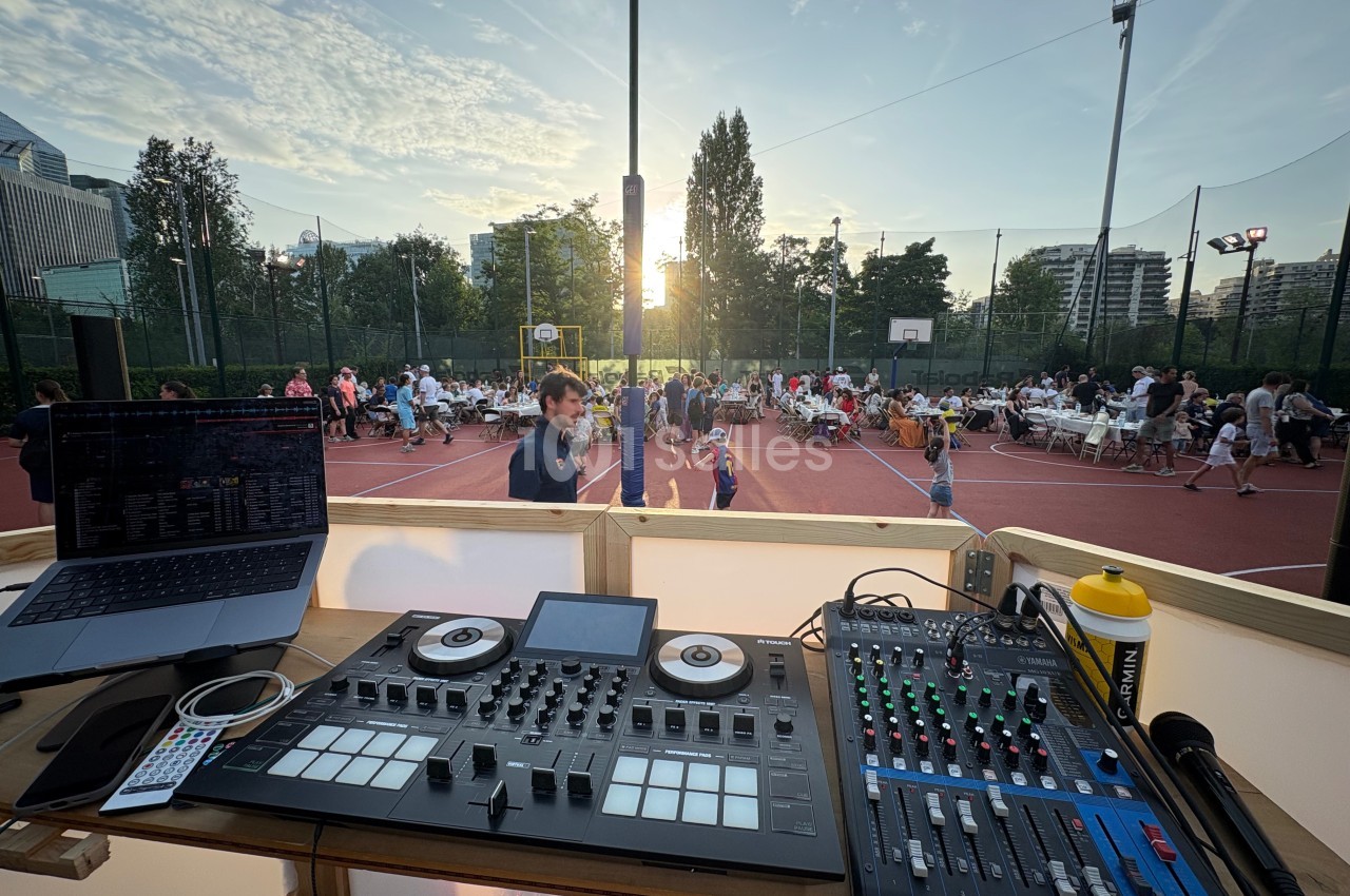 Poste de DJ avec table de mixage, ordinateur et console, surplombant un événement en plein air sur un terrain de sport.