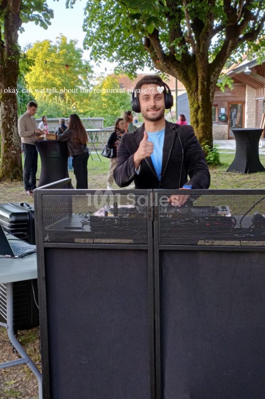 Un DJ souriant derrière une table de mixage en extérieur, avec des invités discutant en arrière-plan.