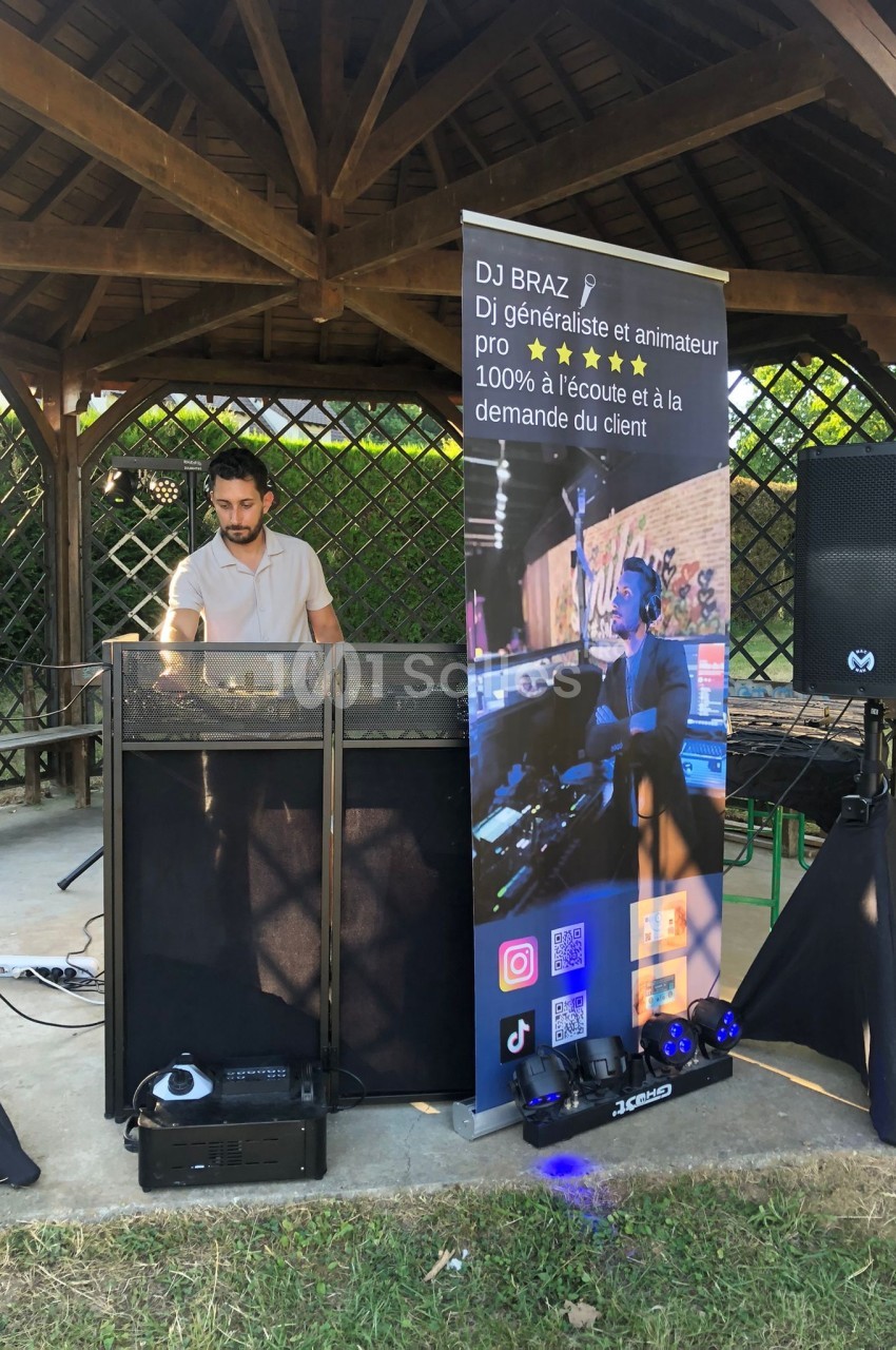 Un DJ mixe sous un kiosque en bois, avec une bannière promotionnelle et du matériel audio installé devant lui.