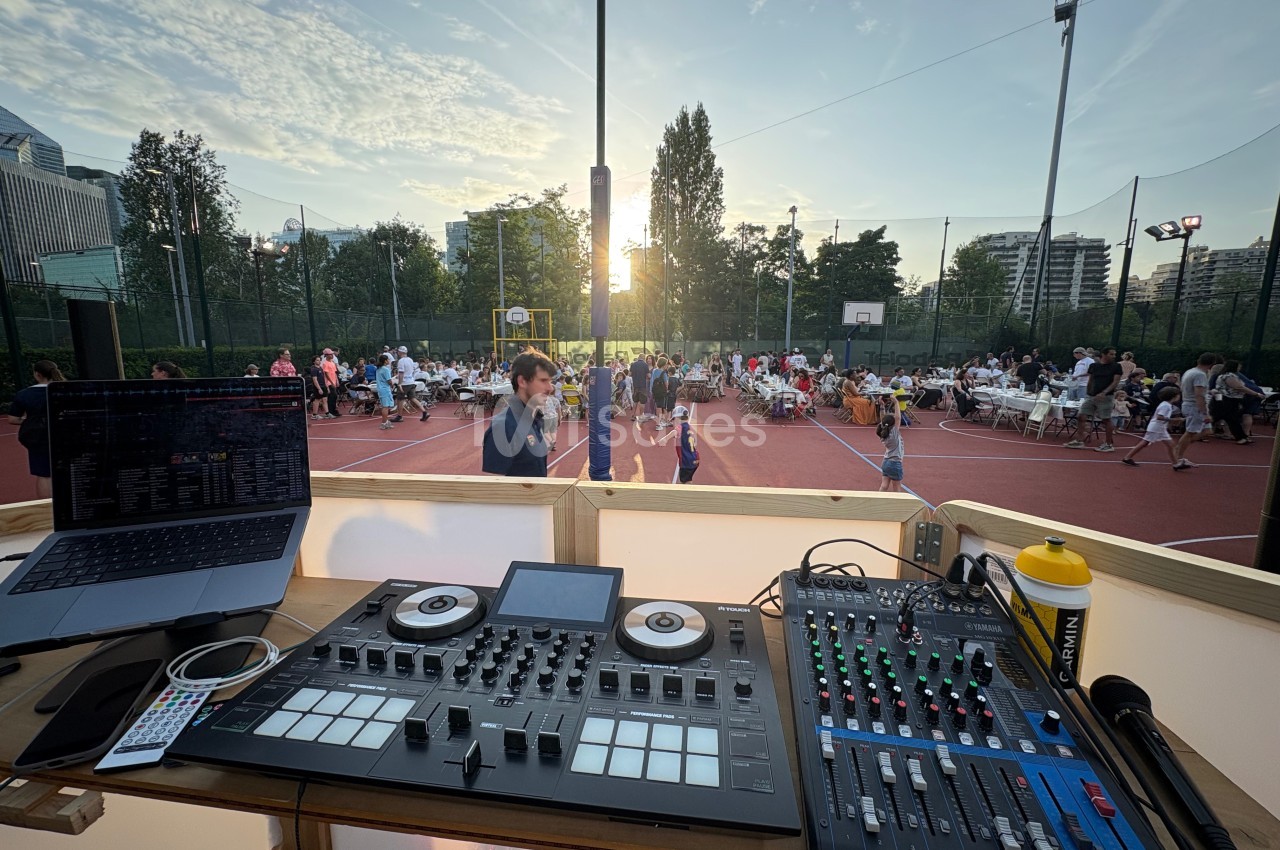 Poste de DJ avec table de mixage, ordinateur et console, surplombant un événement en plein air sur un terrain de sport.