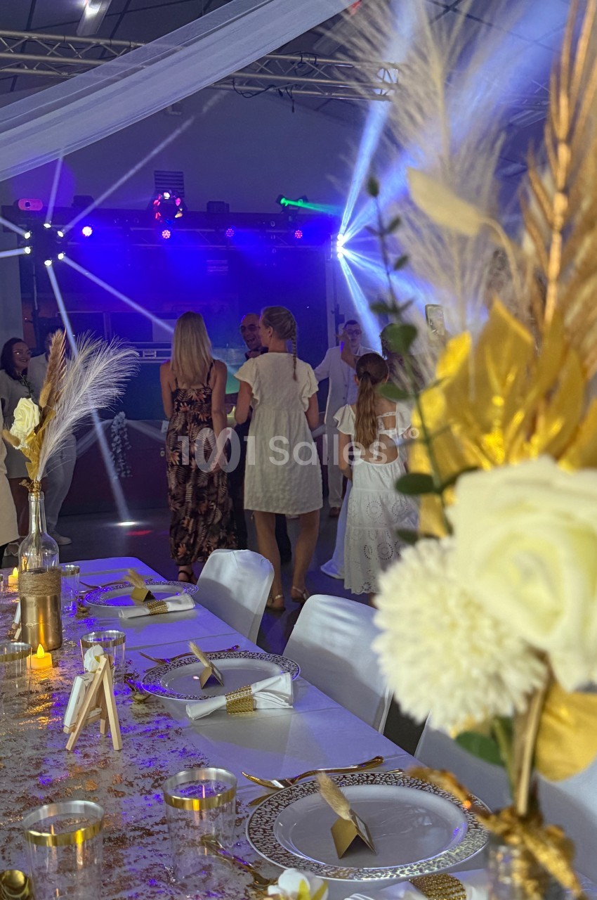 Salle décorée pour un événement avec table élégante au premier plan et invités dansant sous des jeux de lumière en arrière…