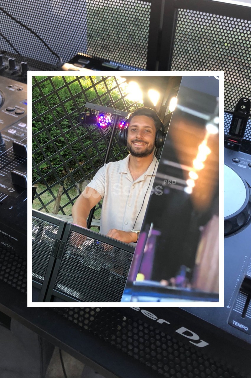 Un homme souriant se tient derrière une table de mixage avec des équipements de DJ en extérieur.